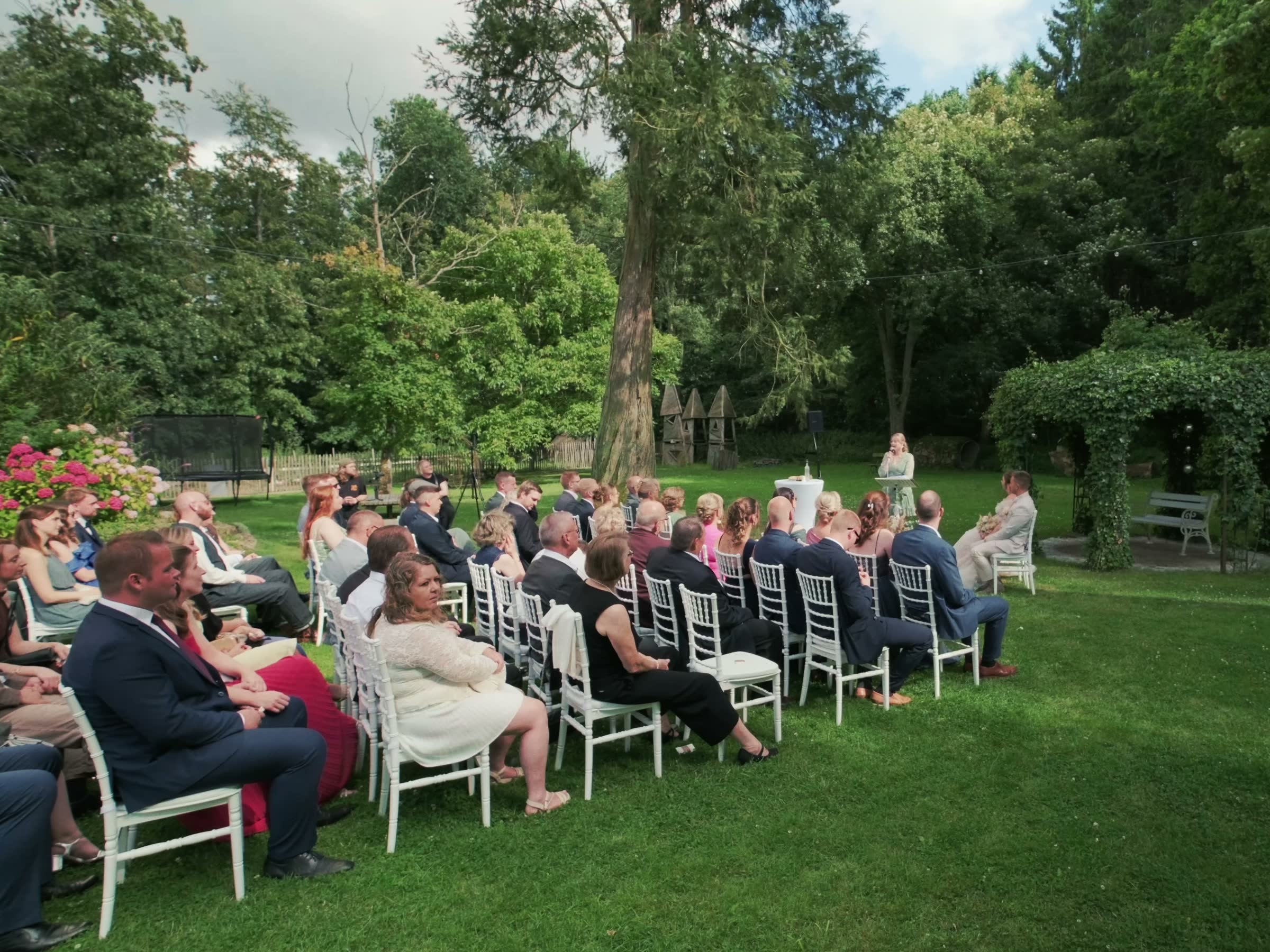 Hochzeitsfilm freie Trauung – Referenz Standbild aus einem Hochzeitsfilm: freie Trauung im Garten mit Gästen auf weißen Stühlen