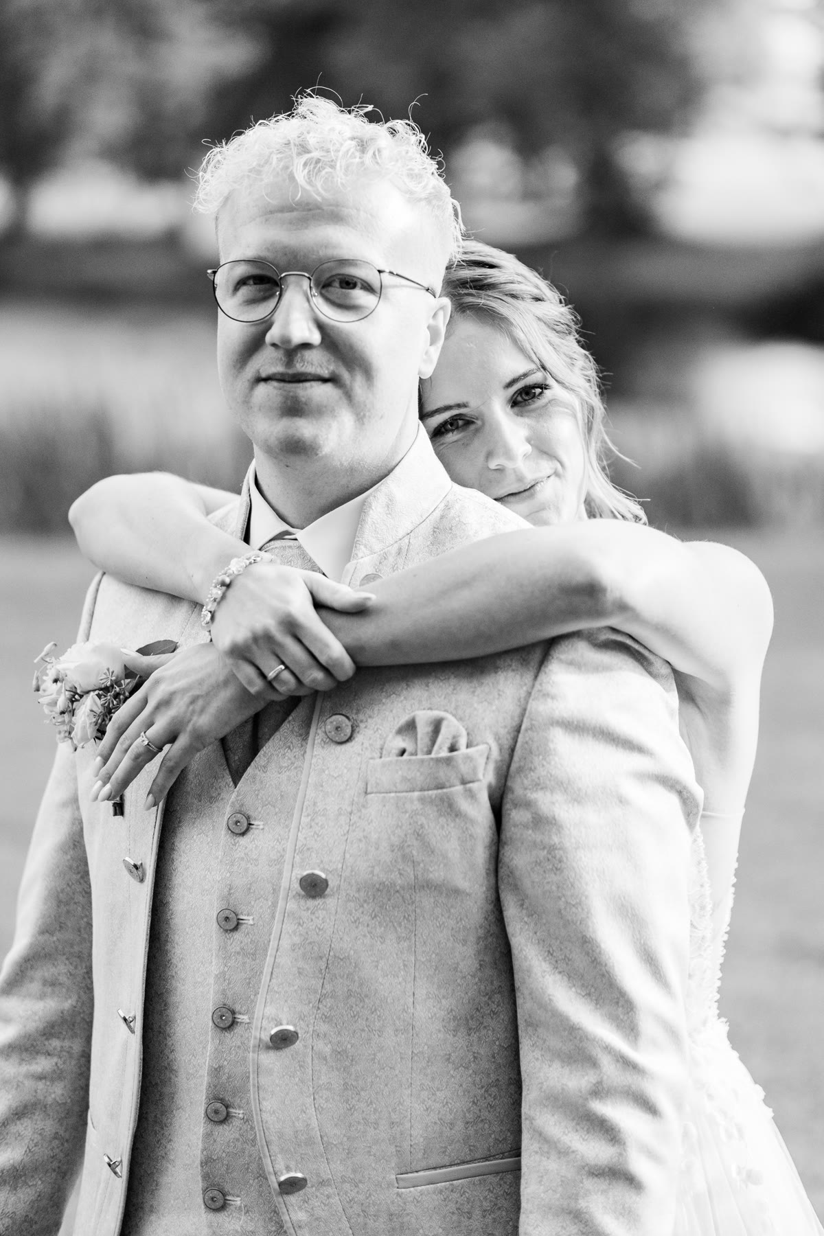 Entspannte Hochzeitsfotografie auch für Kamera-Muffel Bräutigam mit Brille wird lachend von seiner Braut von hinten umarmt, Schwarz-Weiß-Aufnahme